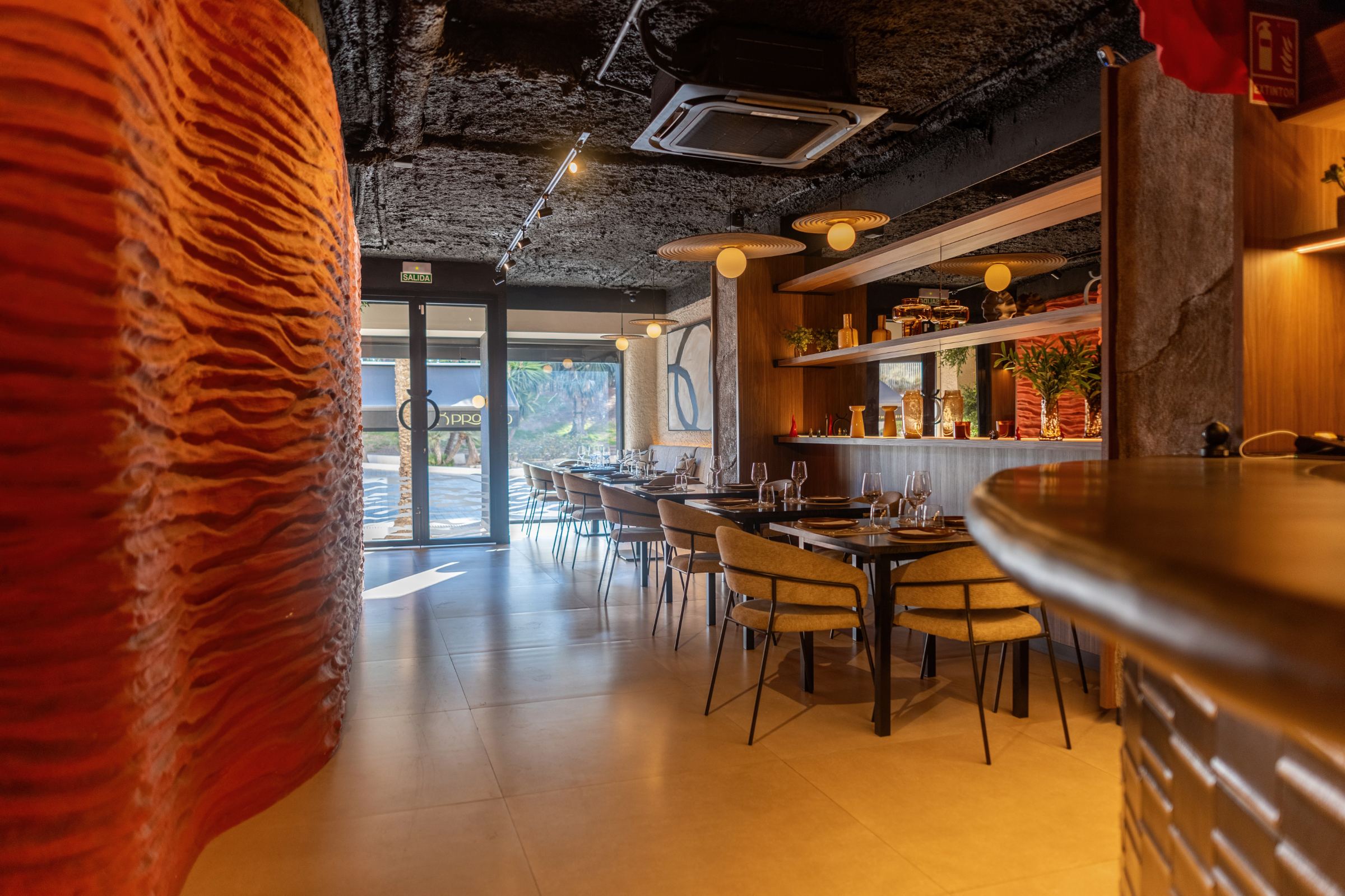 Sala del restaurante Pronto con una gran pared ondulada de color rojo volcánico, barra curva y mesas al fondo