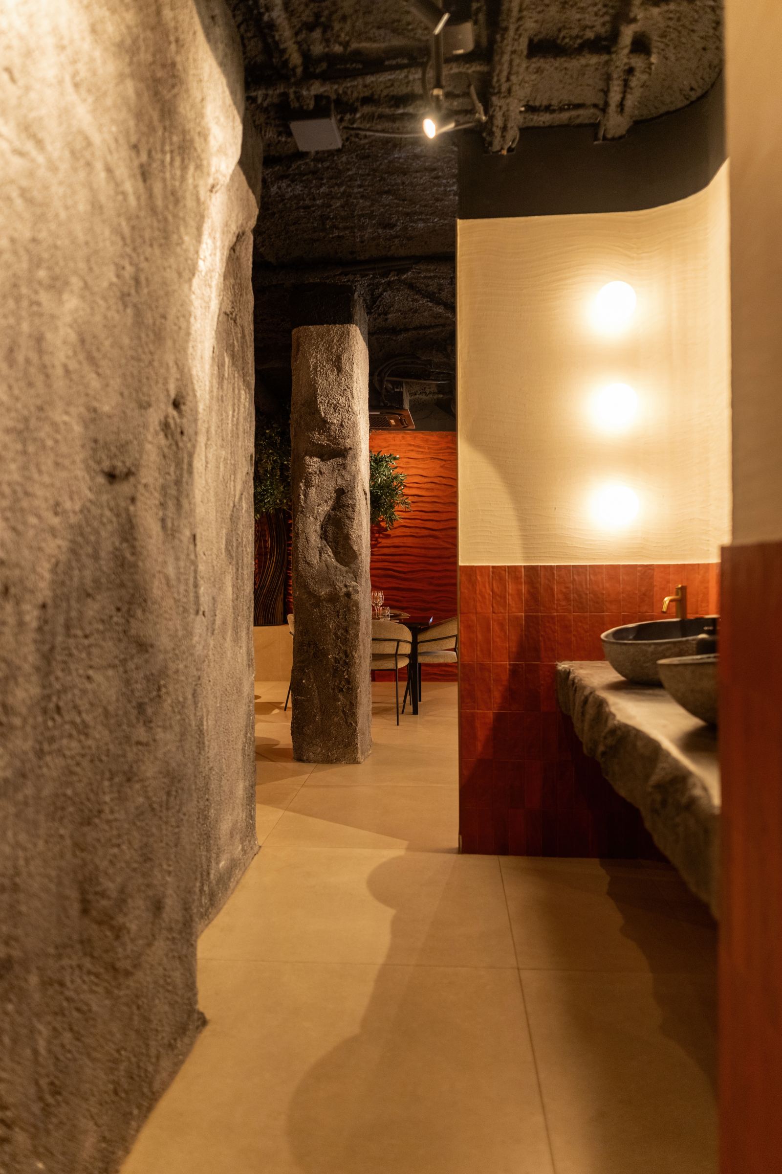 Pasillo de aseos entre muros de piedra, con apliques de luz y lavabo de piedra